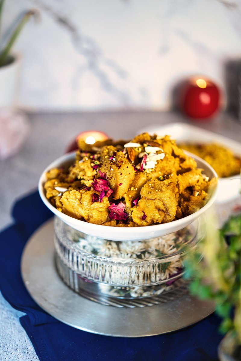 Moong dal halwa served in a decorative bowl on a glass base, garnished with nuts and rose petals. The background features subtle decor and a soft focus environment.