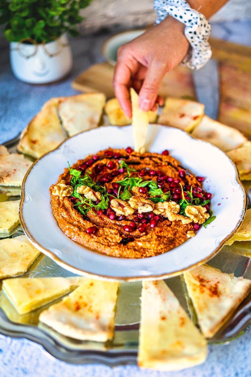 Hand dipping pita bread into authentic Muhammara dip with walnuts, pomegranate seeds, parsley, and olive oil, surrounded by sliced pita bread on a tray