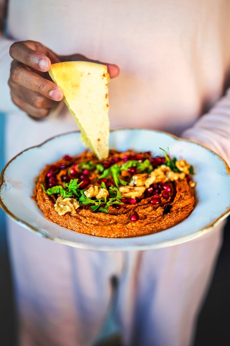 Person holding a plate of homemade Muhammara red pepper walnut dip, garnished with walnuts, parsley, and pomegranate, about to scoop with pita breada