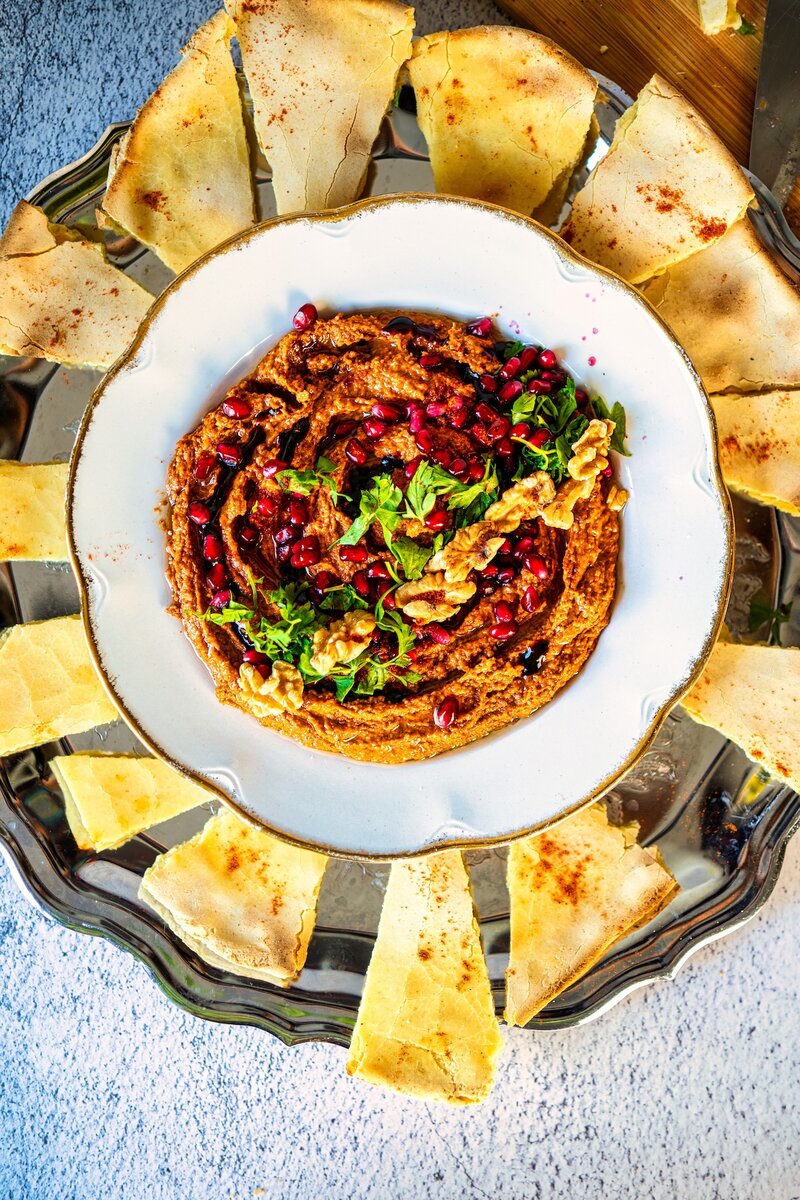 Overhead view of Muhammara dip in a white bowl, beautifully garnished and surrounded by evenly cut pita bread slices on a silver platter