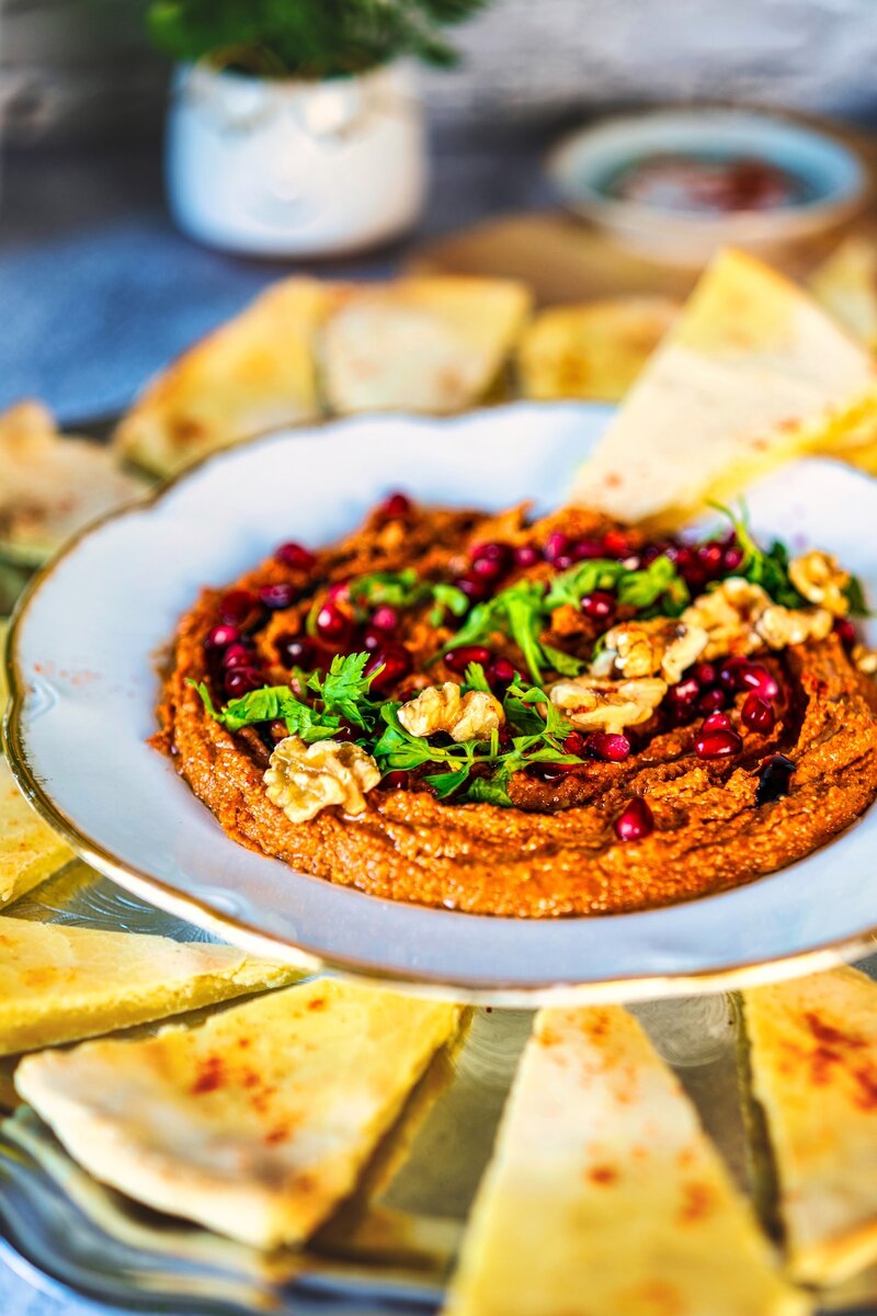 Side angle of vibrant Muhammara dip garnished with walnuts, parsley, and pomegranate arils, arranged with pita slices for serving