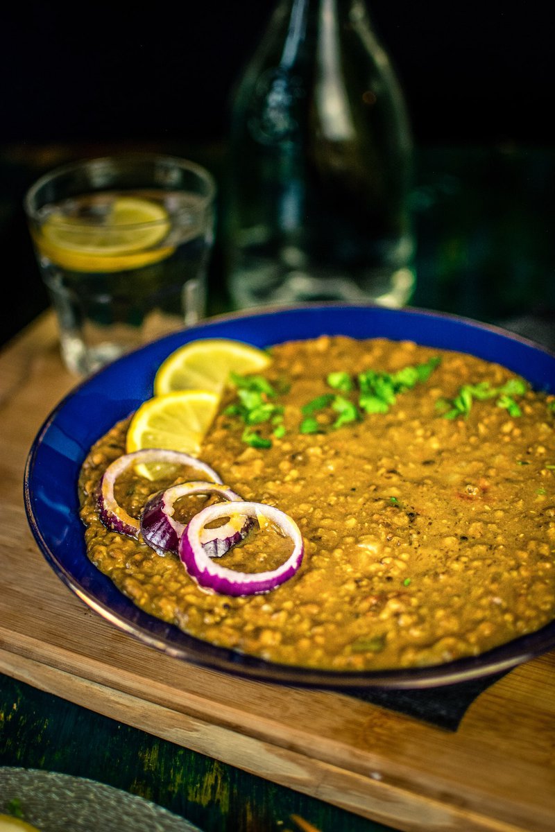 A close-up of a plate of mung bean curry garnished with lemon slices and red onion rings, with a blurred glass of water and bottle in the background.