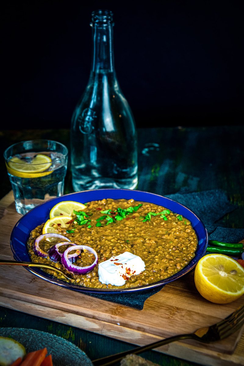 Instant pot mung bean curry or whole green moong dal plated with a dollop of yogurt and a sprinkle of spice on top, accompanied by lemon wedges and onion rings, with a glass of water and bottle in soft focus behind.