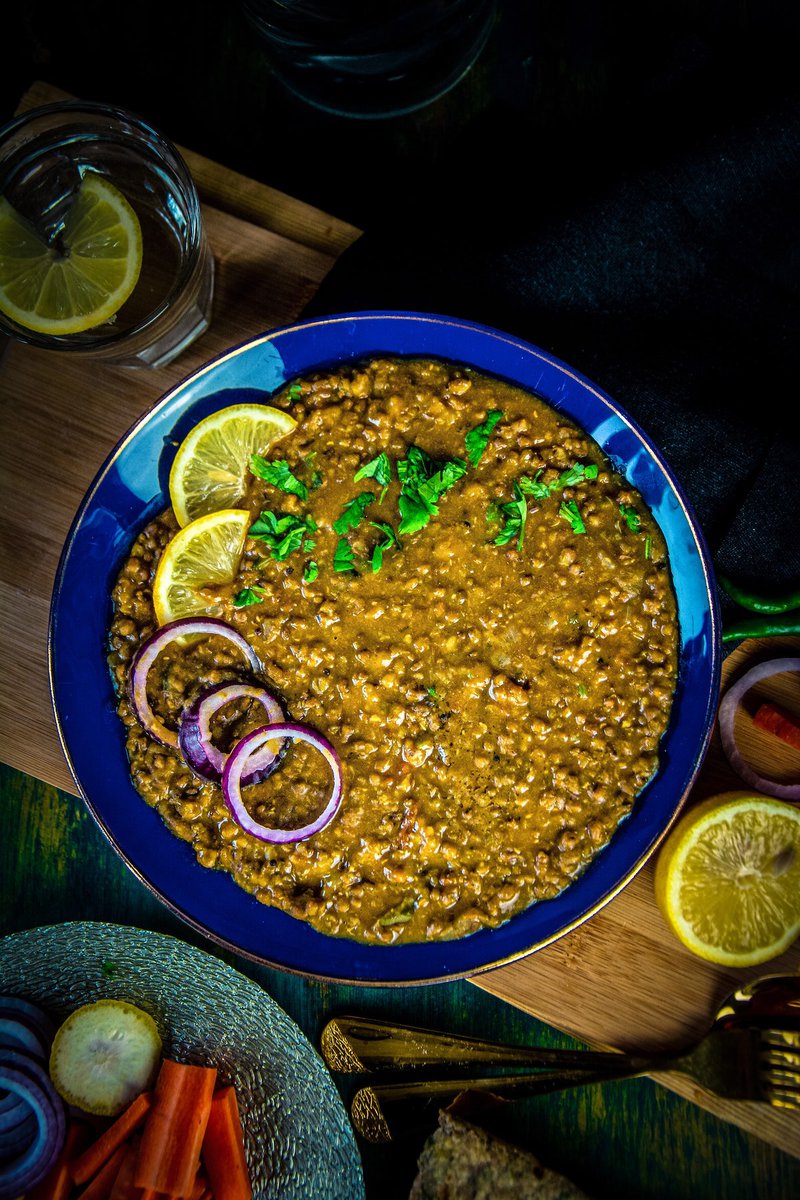 Overhead shot of a vibrant bowl of green whole moong dal or mung bean curry topped with fresh herbs, next to cut vegetables and a piece of flatbread, on a dark wooden background.