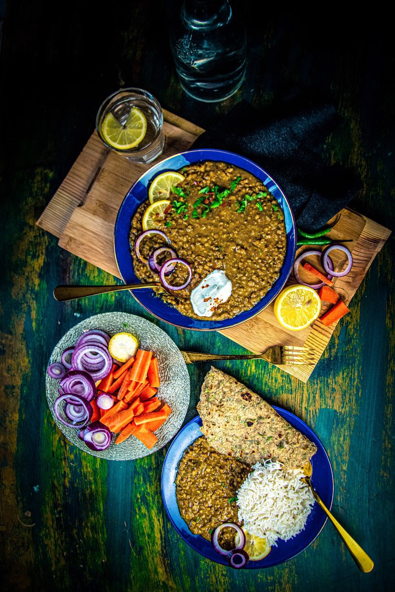 An appetizing arrangement featuring a bowl of mung bean curry aka whole green moong, alongside rice, flatbread, and a small bowl of cut vegetables, on a wooden board.
