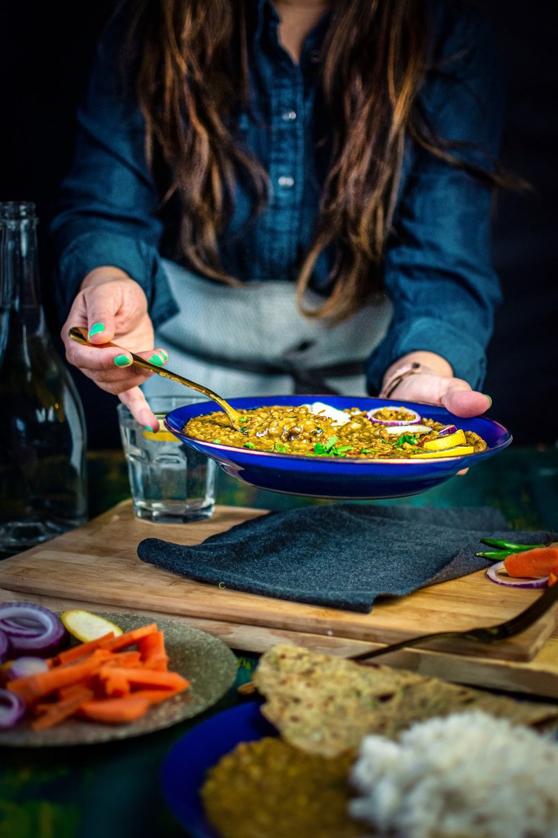 A bowl of instant pot mung bean curry held by a person with a denim shirt, with a wooden table setting including a water glass, vegetables, and flatbread.