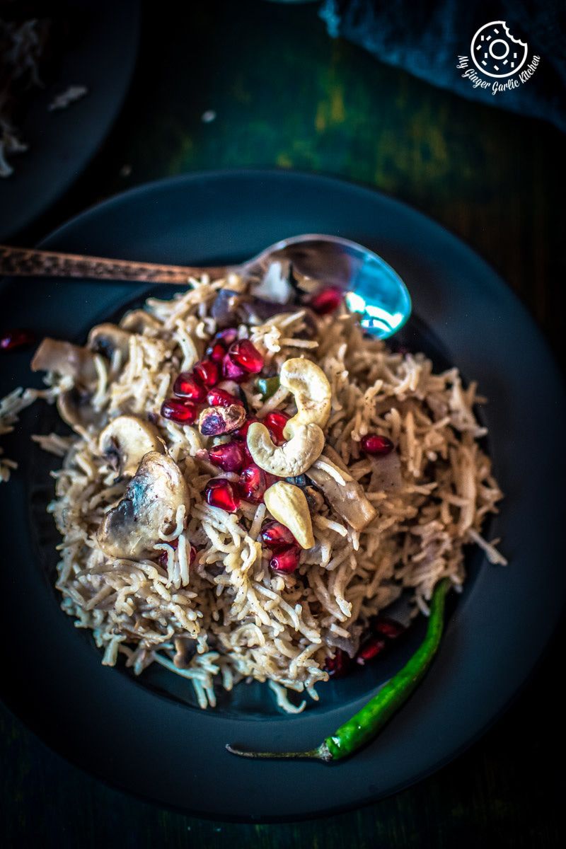 a plate of mushroom pulao topped with cashews, and pomegranate seeds with a spoon on it