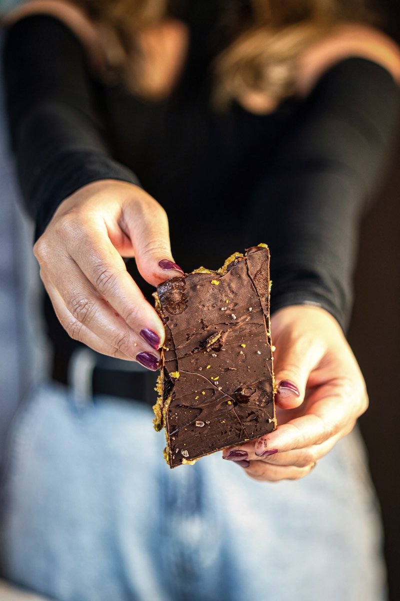 Homemade no-bake almond energy bar held in both hands, showing the chewy nut and dried fruit filling under the chocolate topping.