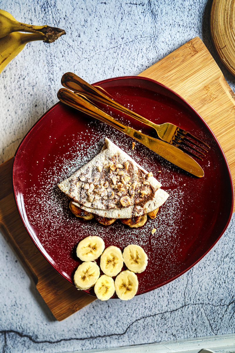 Overhead view of a nutella banana wrap with banana slices and nuts on a red plate, with gold utensils.