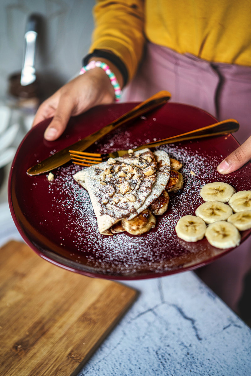 Hands holding a burgundy plate with a nutella banana wrap filled with banana and nuts, dusted with powdered sugar.