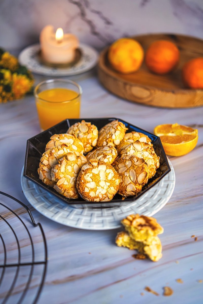 Orange almond cookies topped with sliced almonds, served on a black plate with fresh orange juice and oranges