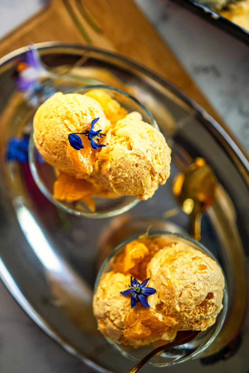 Two scoops of orange ice cream in a glass bowl garnished with blue flowers, served on a reflective silver tray.