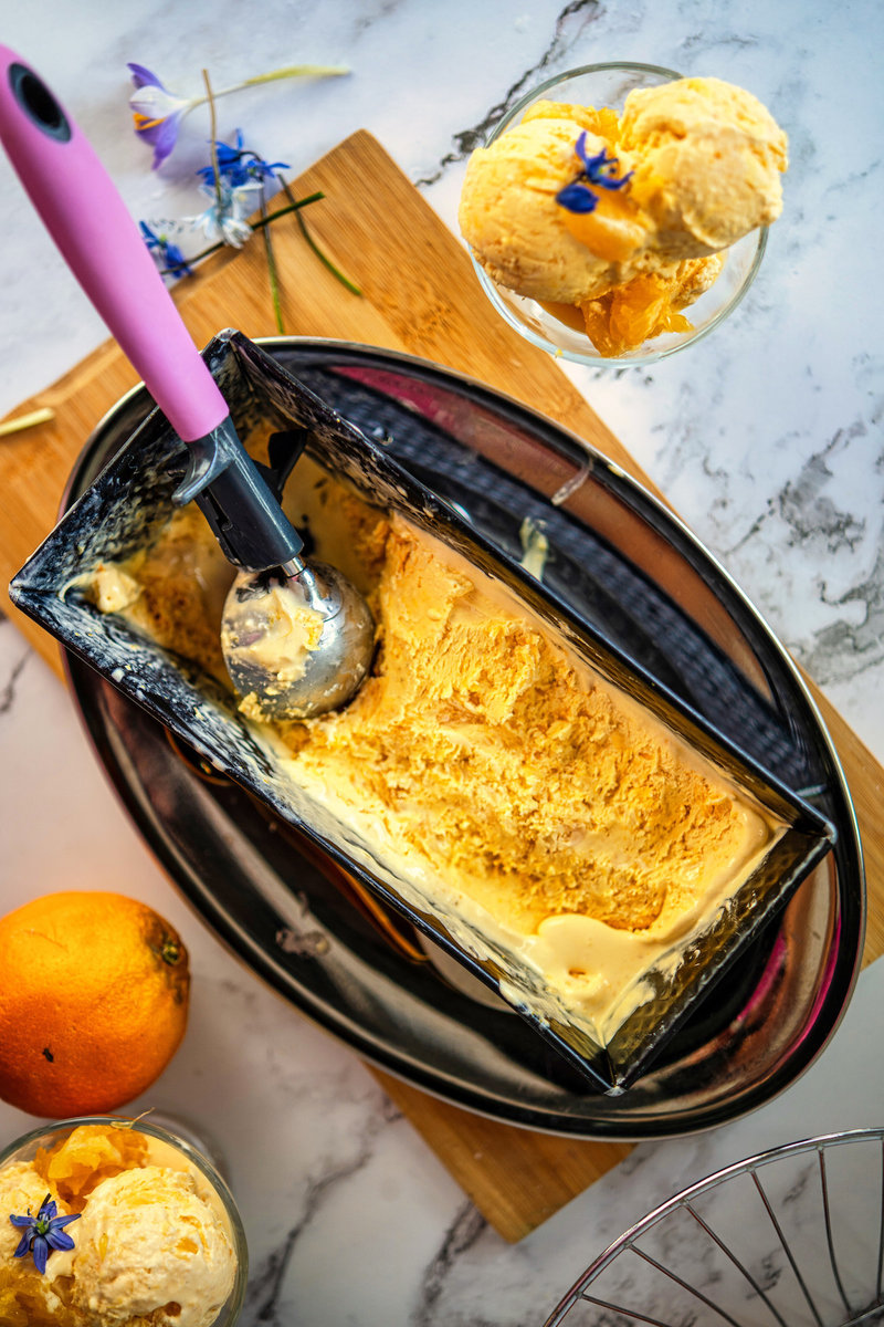 A loaf pan filled with orange ice cream beside a scoop, with whole oranges and blue flowers on a marble surface.