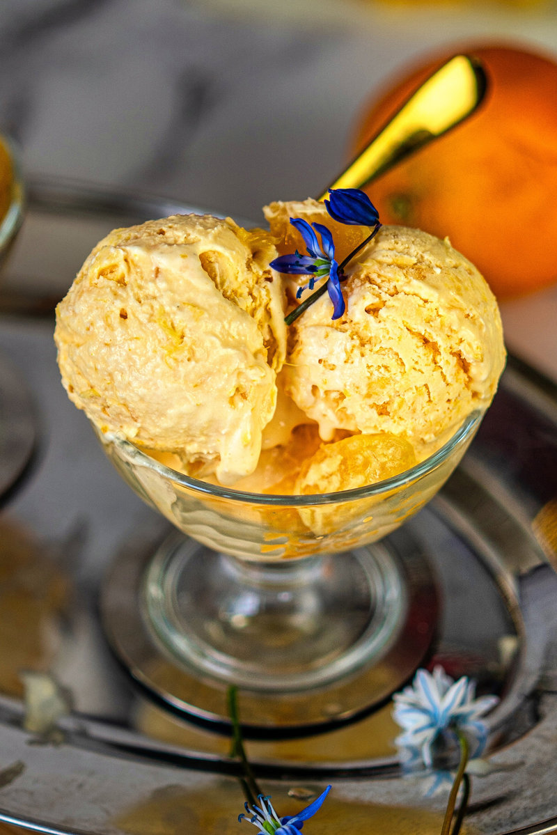 Close-up of two scoops of orange ice cream in a glass bowl with a blue flower garnish, showing creamy texture.