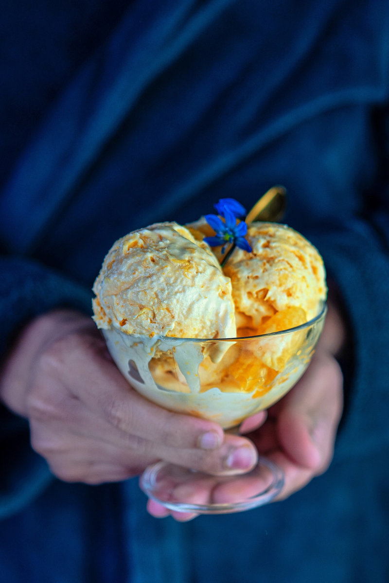 Hand holding a glass bowl filled with orange ice cream scoops, garnished with a blue flower, with a blurred background.