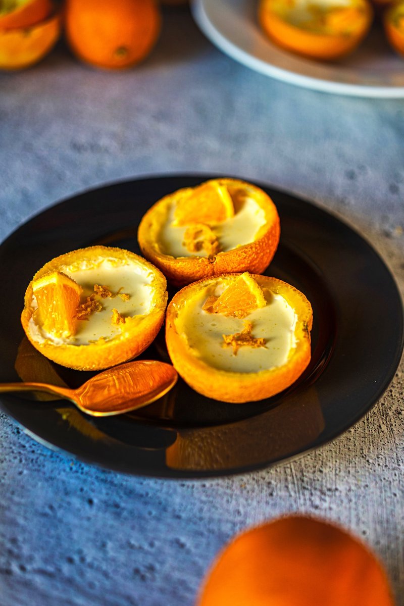 Creamy orange posset served in fresh orange shells on a black plate, garnished with orange zest and juicy orange wedges.