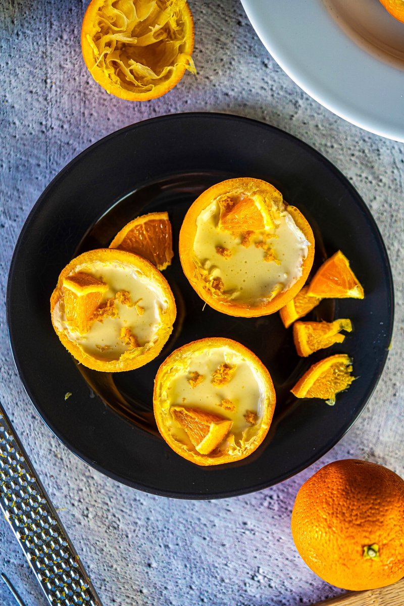 Overhead shot of easy orange posset recipe served in orange peels with fresh citrus garnish on a black serving plate.