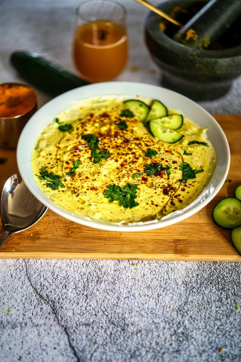 Pahadi kheera ka raita on a white plate topped with chopped cilantro, spices, cucumber slices, and flatbread on a wooden board with a juice glass in the background.