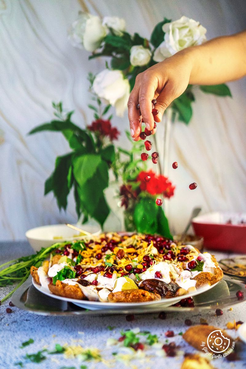 a hand sprinkling pomegranate arils over pakora chaat plate