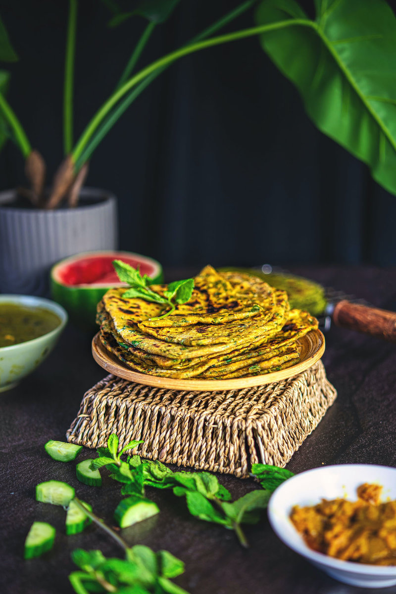 Stack of homemade palak paratha aka spinach paratha on a wooden plate with fresh mint leaves on the side, atop a rustic woven stand.