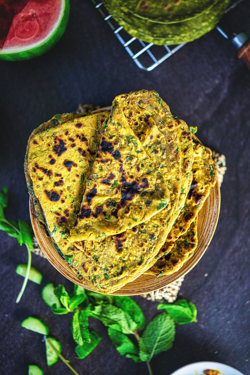 Overhead view of palak paratha aka spinach flabread on a wooden plate, garnished with mint leaves, with a backdrop of vibrant watermelon and greenery.