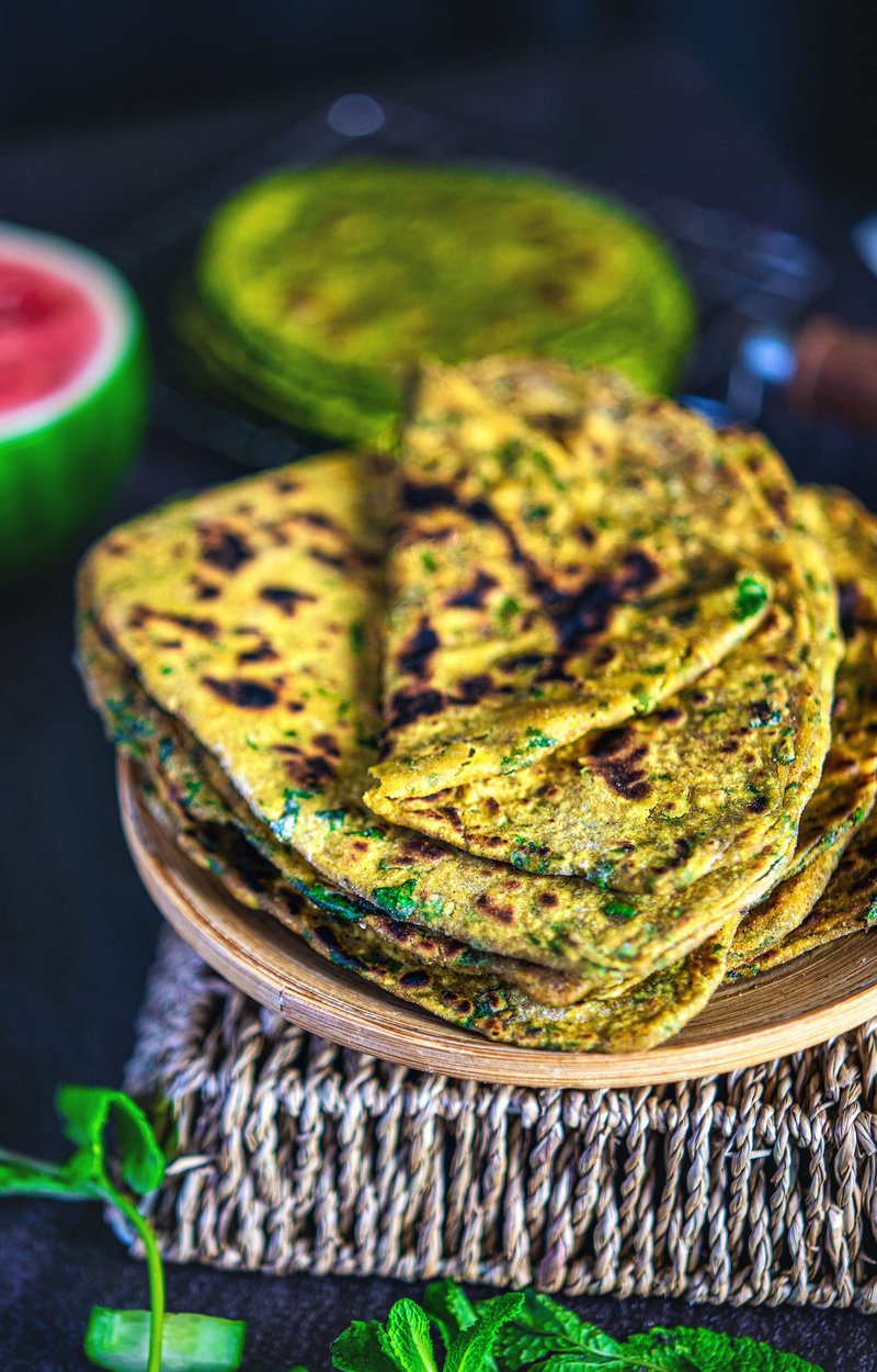 Close-up of golden-brown palak paratha on a wooden plate, surrounded by fresh mint leaves on a dark textured surface.
