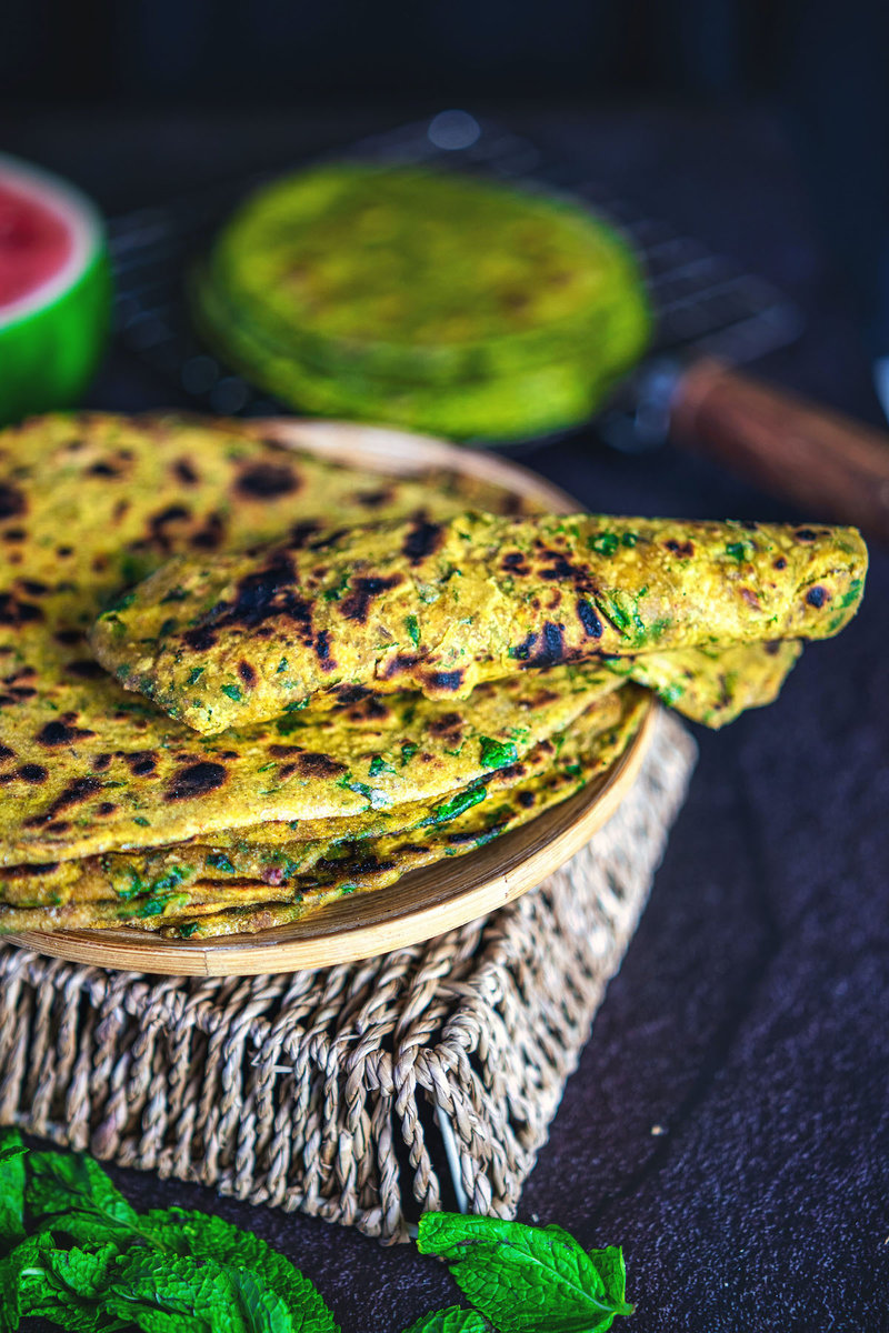 Angled view of a pile of palak paratha on a plate, with a piece folded to reveal the inner spinach layers, accompanied by mint leaves.
