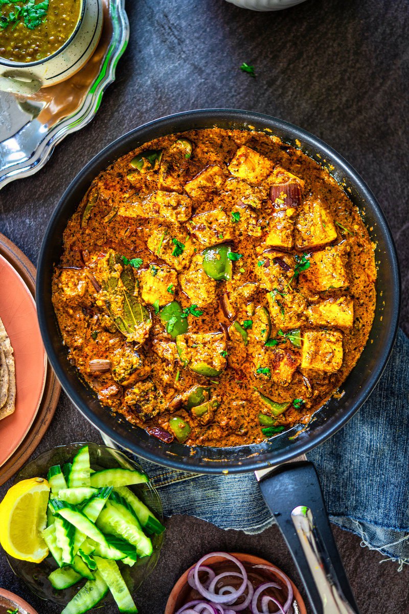 Top view of paneer bhuna masala in a pan, garnished with green bell peppers and cilantro, accompanied by a bowl of dal and fresh vegetables on a dark table.