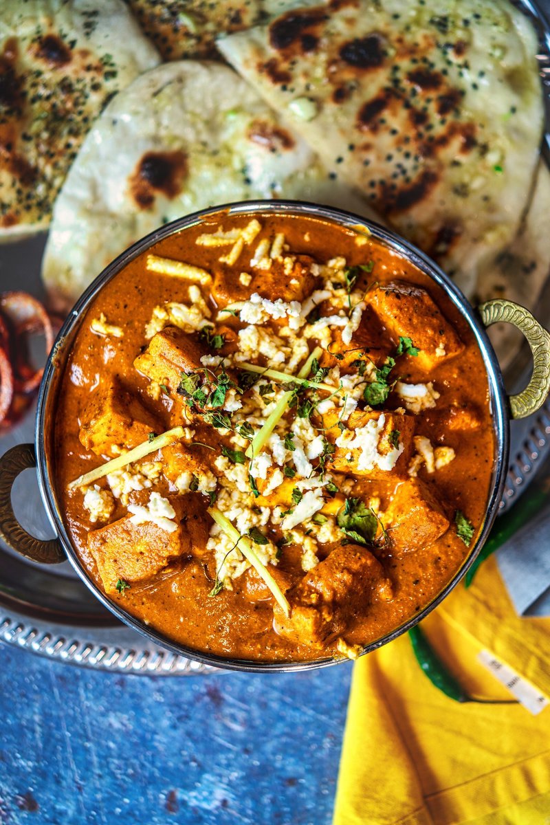 A vibrant bowl of paneer lababdar, topped with crumbled paneer and julienned ginger, accompanied by charred naan bread, on a patterned silver tray with a yellow napkin.