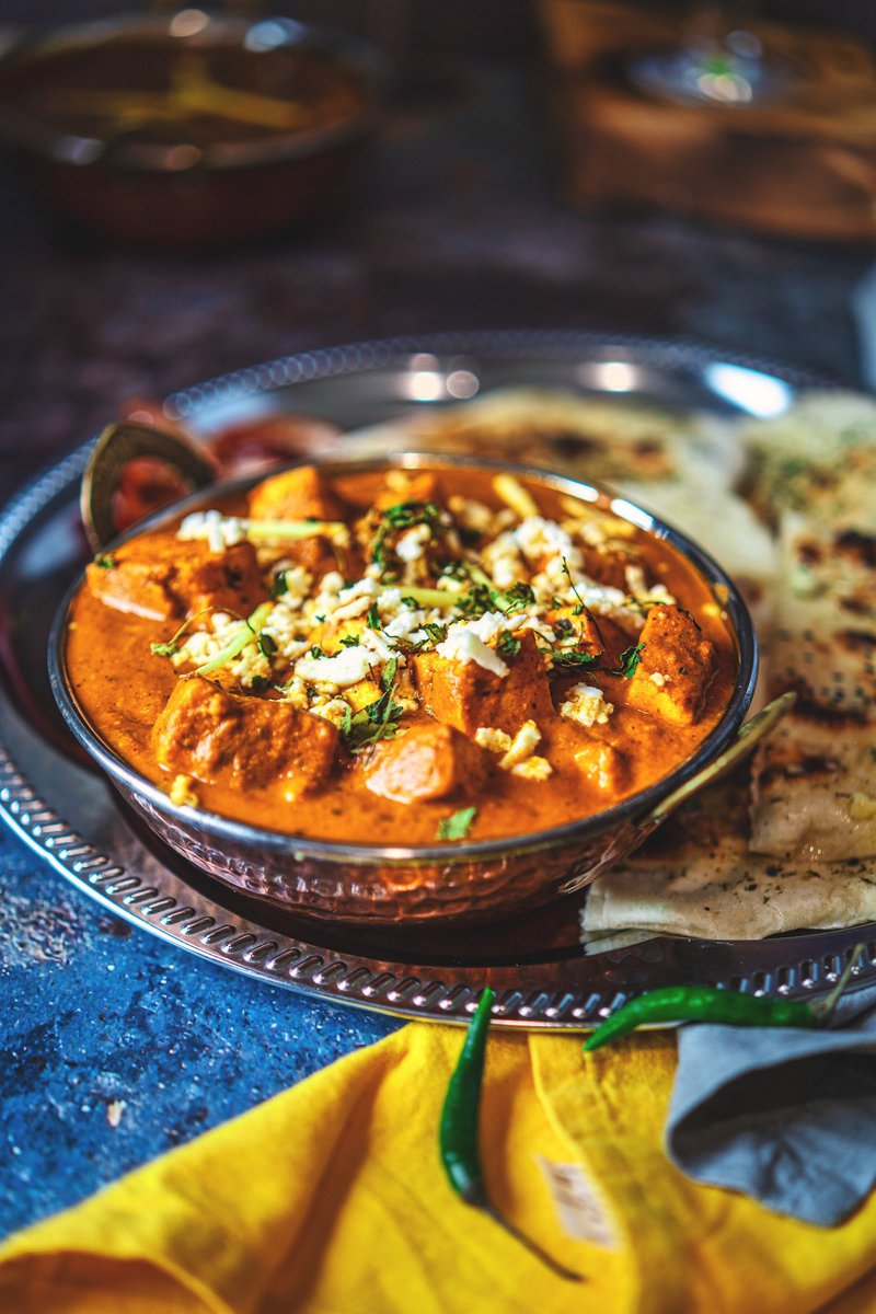 A serving of paneer lababdar in a copper bowl on a blue textured surface, accompanied by various Indian breads and a side dish in the blurred background.