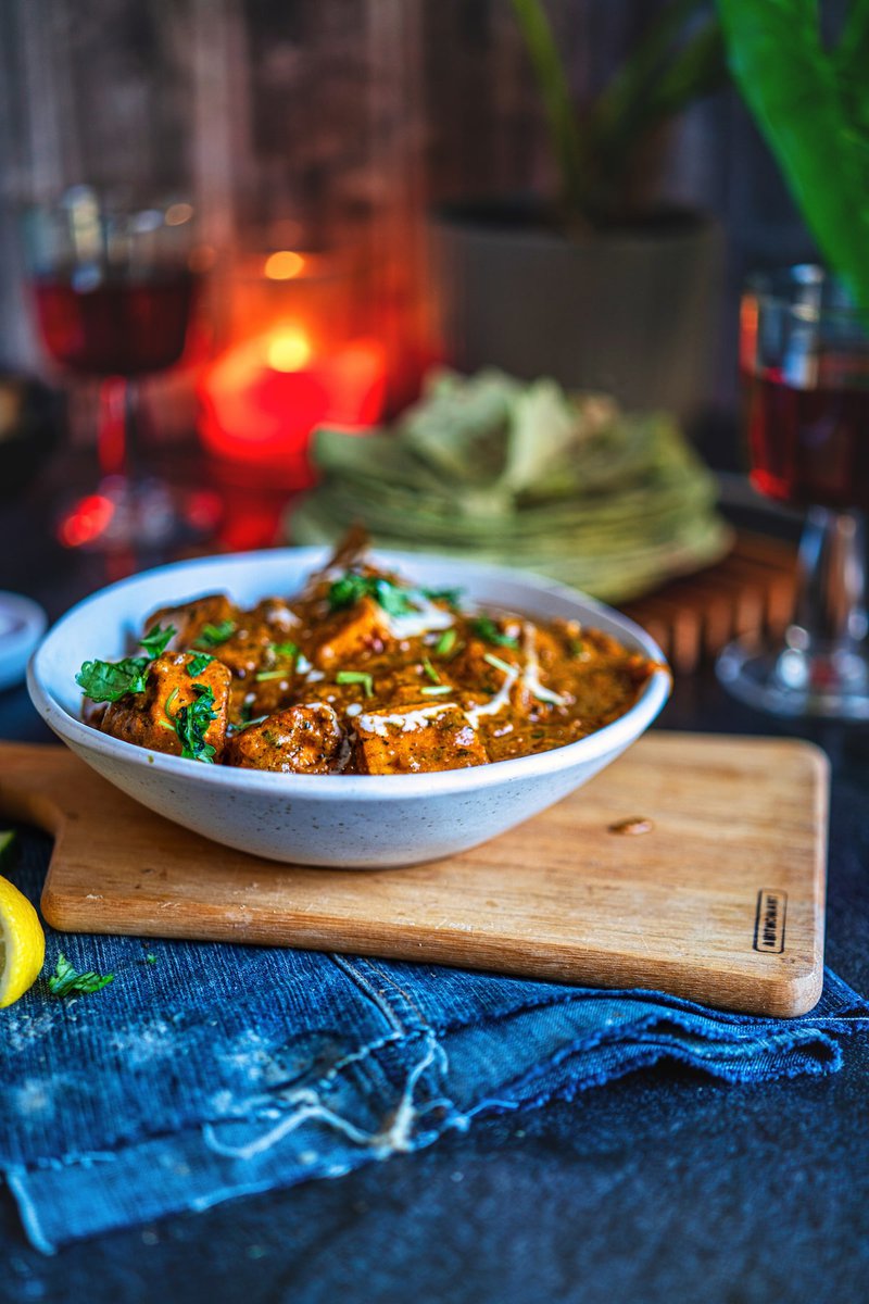 A close-up of a bowl filled with paneer masala, garnished with coriander and drizzled with cream. The background shows a candle-lit table setting, adding a cozy ambiance with a glass of wine and folded chapatis