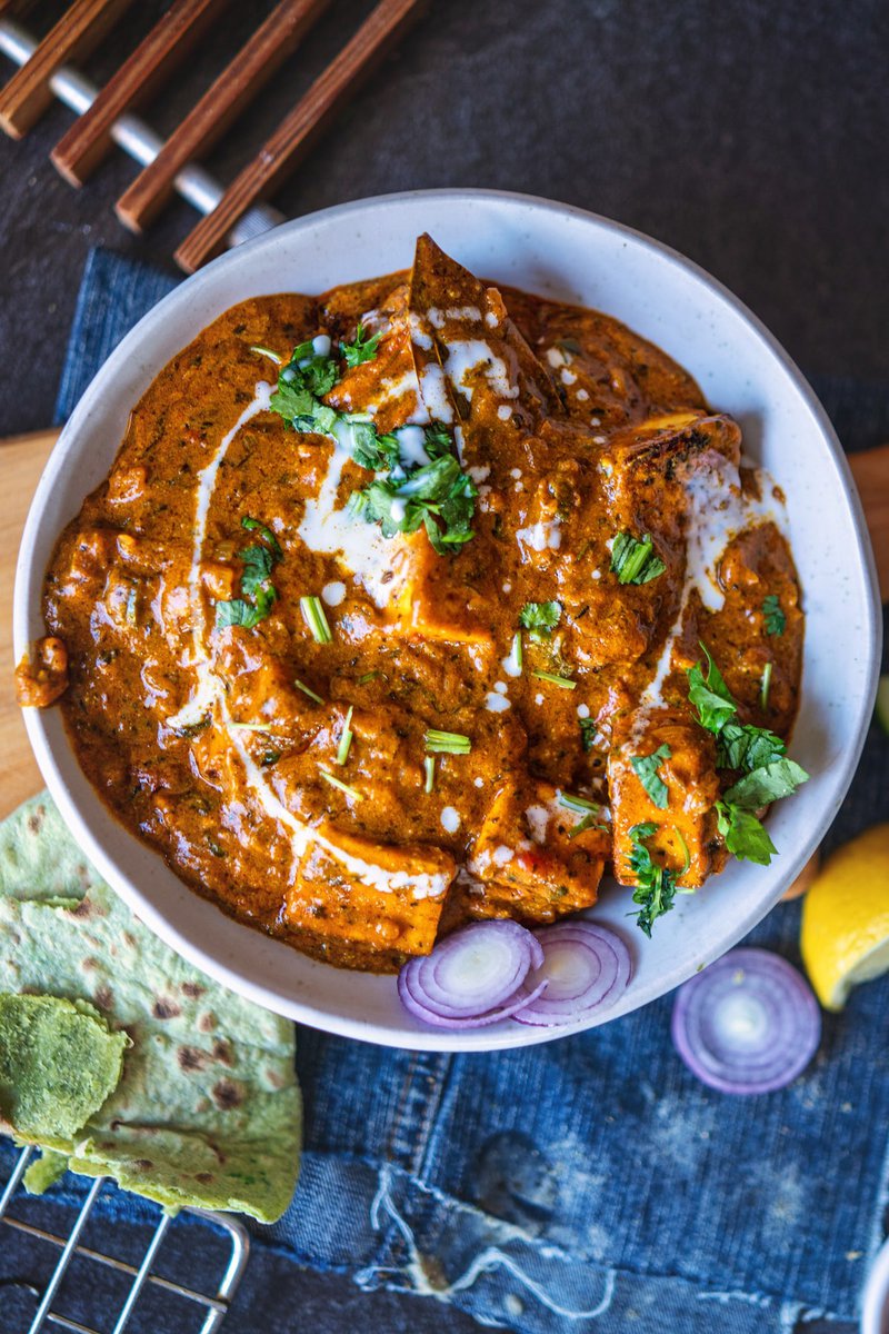 A top-down view of a bowl of paneer masala, with chunks of paneer in a rich, creamy curry sauce, garnished with fresh coriander and cream. The side of the plate has slices of onion and a lemon wedge