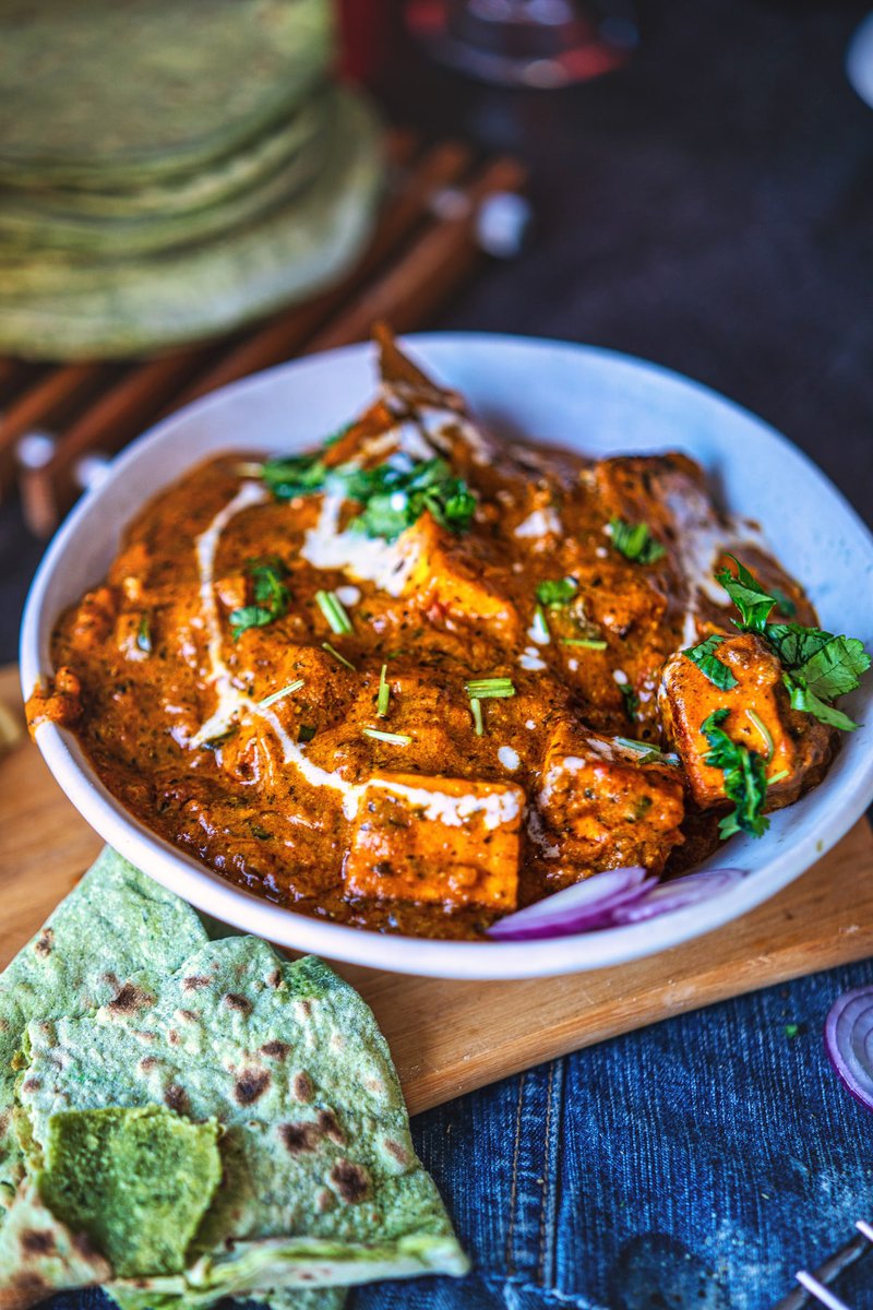 Bowl of paneer masala curry with cubes of paneer cheese in a rich, orange-colored sauce garnished with fresh cilantro. Served alongside green flatbread on a wooden board