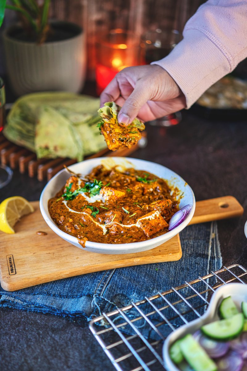 A hand dipping a piece of chapati into a bowl of paneer masala, with pieces of paneer in a thick curry sauce, garnished with coriander. The setting is warm with candles, chapatis stacked in the background, and other food accompaniments like cucumber salad