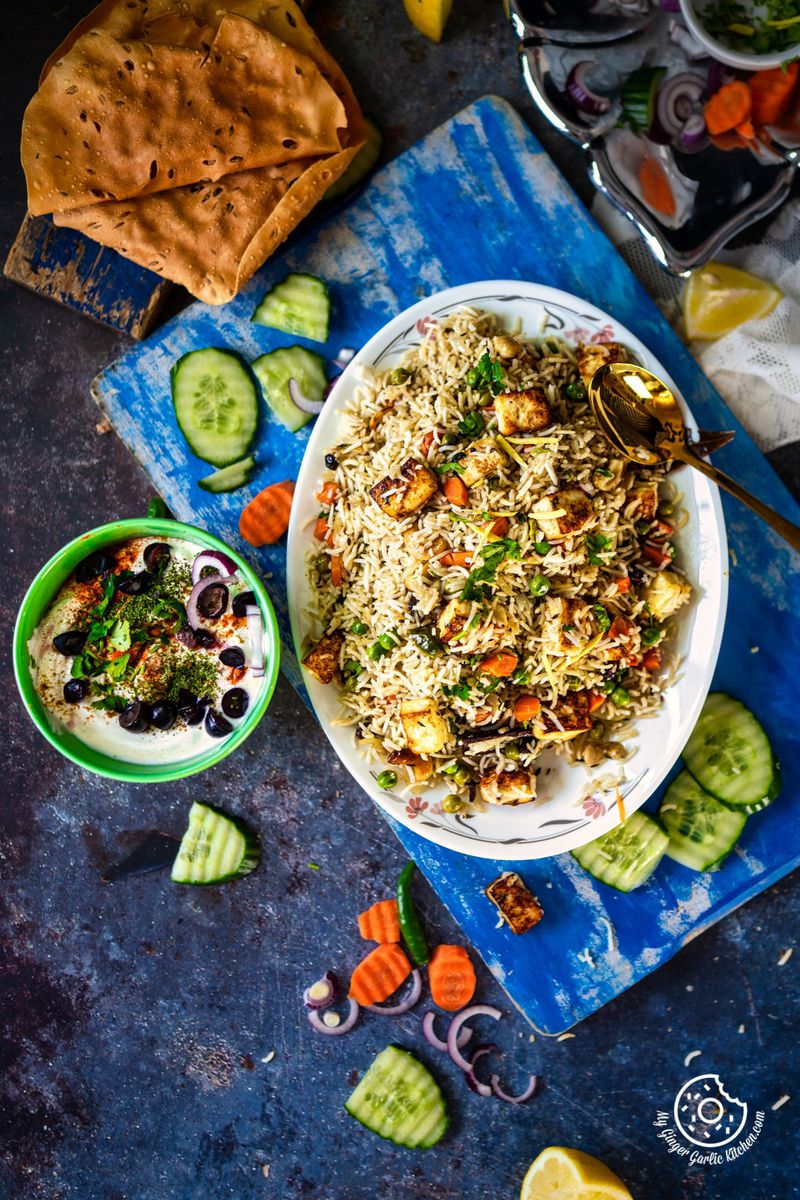 overhead shot of paneer pulao served on a white plate with a side of raita and some papads