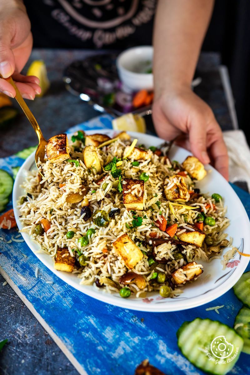 a hand holding paneer pulao plate with one hand and golden spoon with other hand