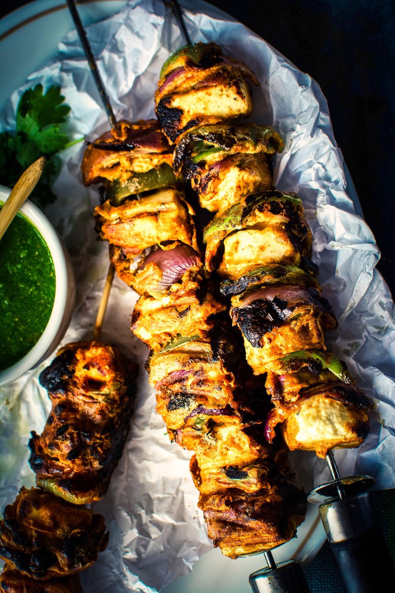 Charred paneer tikka skewers served with a side of green chutney on a white plate, with a moody dark backdrop.