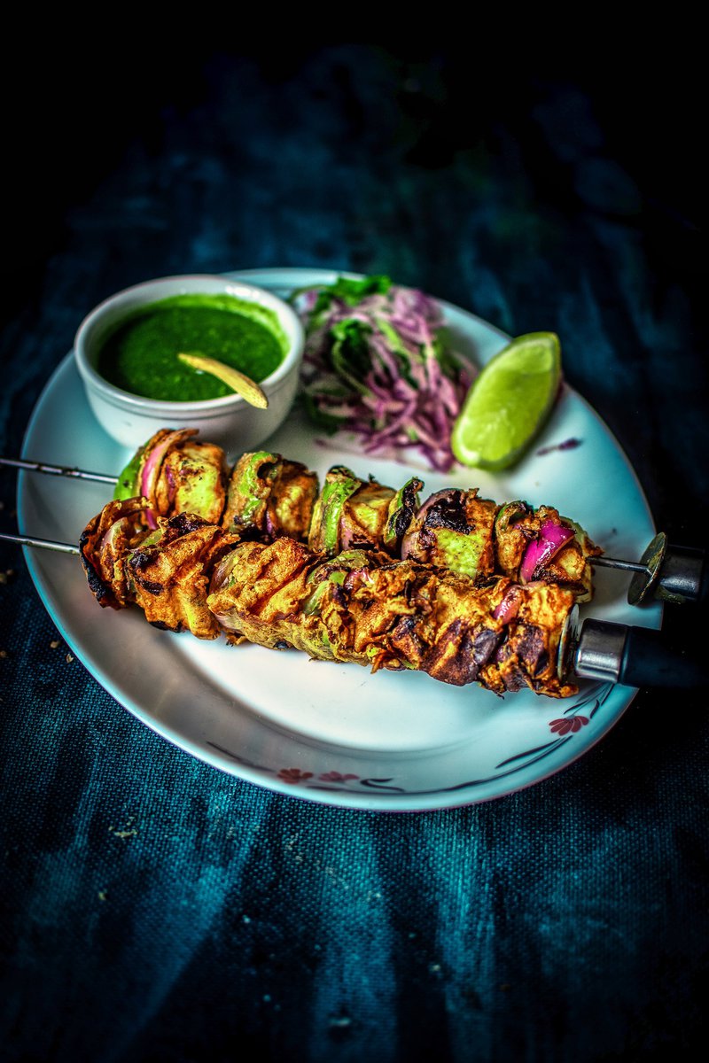  colorful and inviting shot of paneer tikka skewers on a plate with green chutney and salad, against a dark blue backdrop.
