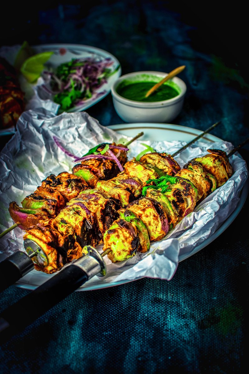 A close-up of succulent paneer tikka skewers, charred and golden, with layers of onion and bell pepper, presented on a white plate with crinkled white paper.