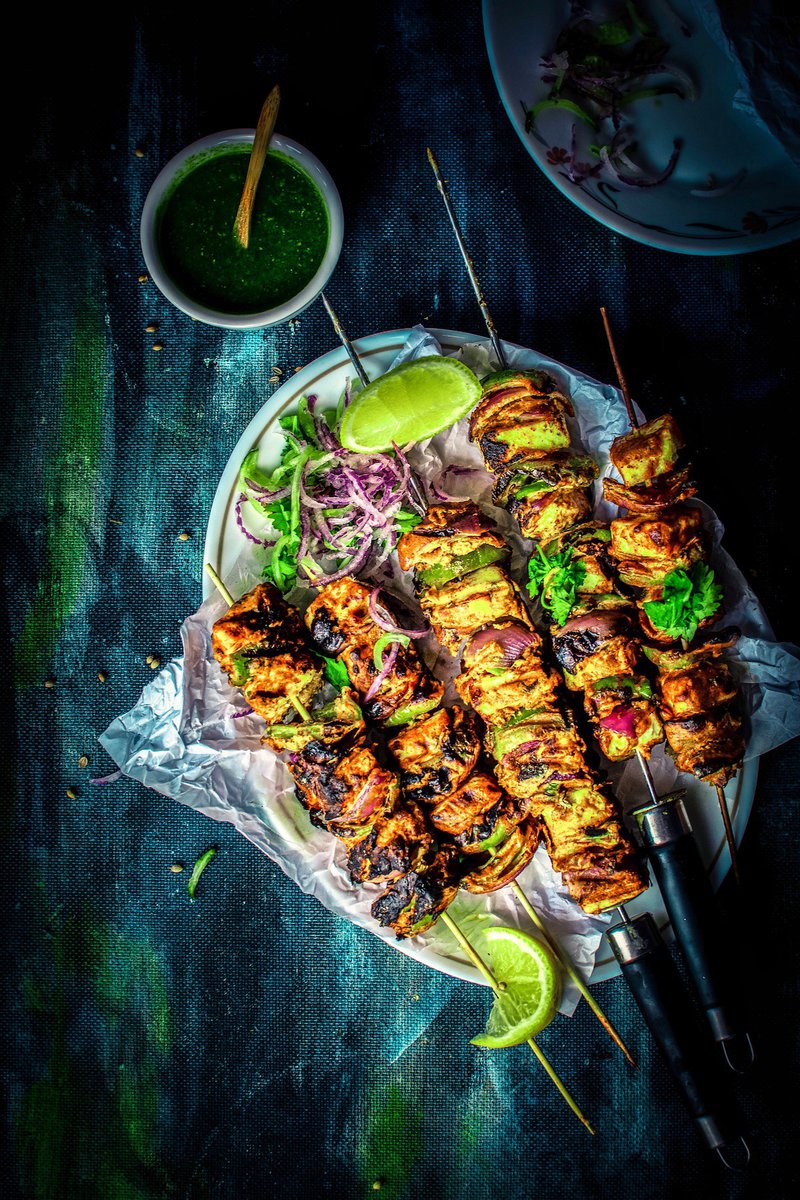 Overhead shot of tandoori paneer tikka skewers with a bowl of green chutney and a salad, on a rustic surface.
