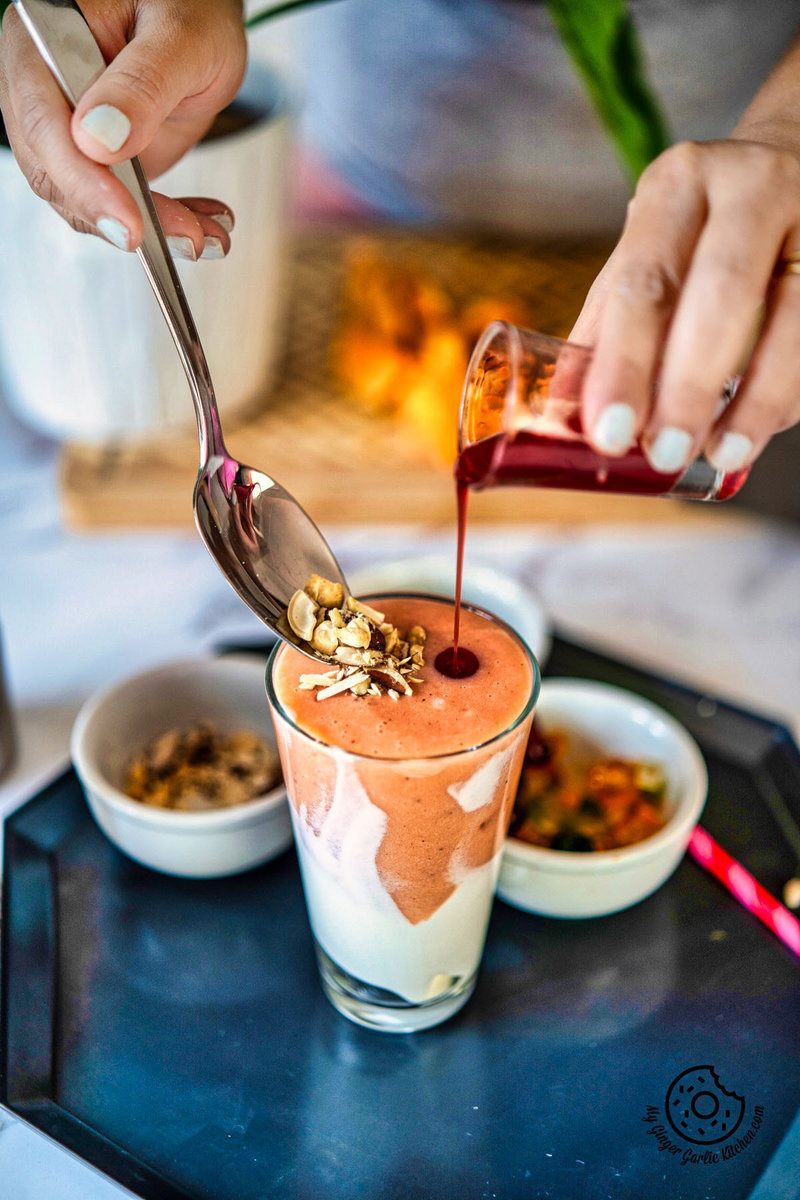 rose syrup being poured with one hand and other hand holding a spoon with nuts on a papaya shake glass