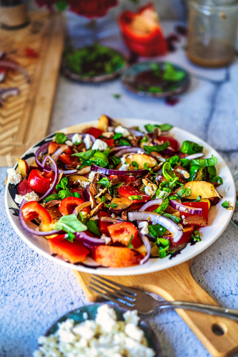 Fresh peach salad with vibrant layers of sliced peaches, red onions, and bell peppers, sprinkled with green basil, goat cheese, and almonds, served on a rustic wooden board.