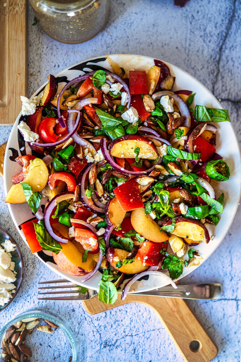 overhead shot of colorful peach salad displayed on a white plate, featuring layers of juicy peach slices, diced red peppers, purple onions, fresh green herbs, and crumbled cheese.