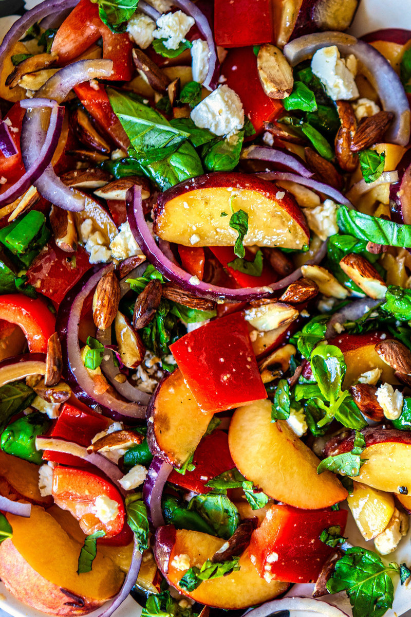 Close-up of a peach salad showcasing bright peach slices mixed with red bell peppers, red onions, and a sprinkle of green herbs and nuts.