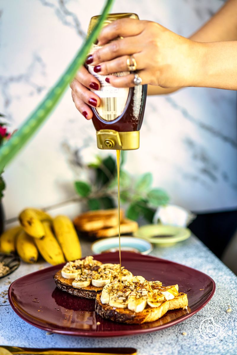 someone pouring honey onto a plate of two peanut butter banana toasts on a table