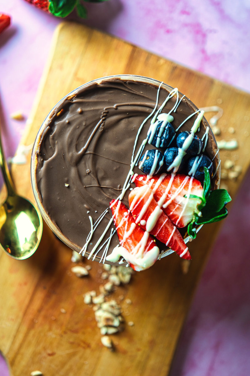 A top-down view of a decadent peanut butter yogurt bowl topped with a chocolate drizzle, fresh strawberries, and blueberries on a wooden board with scattered nuts.