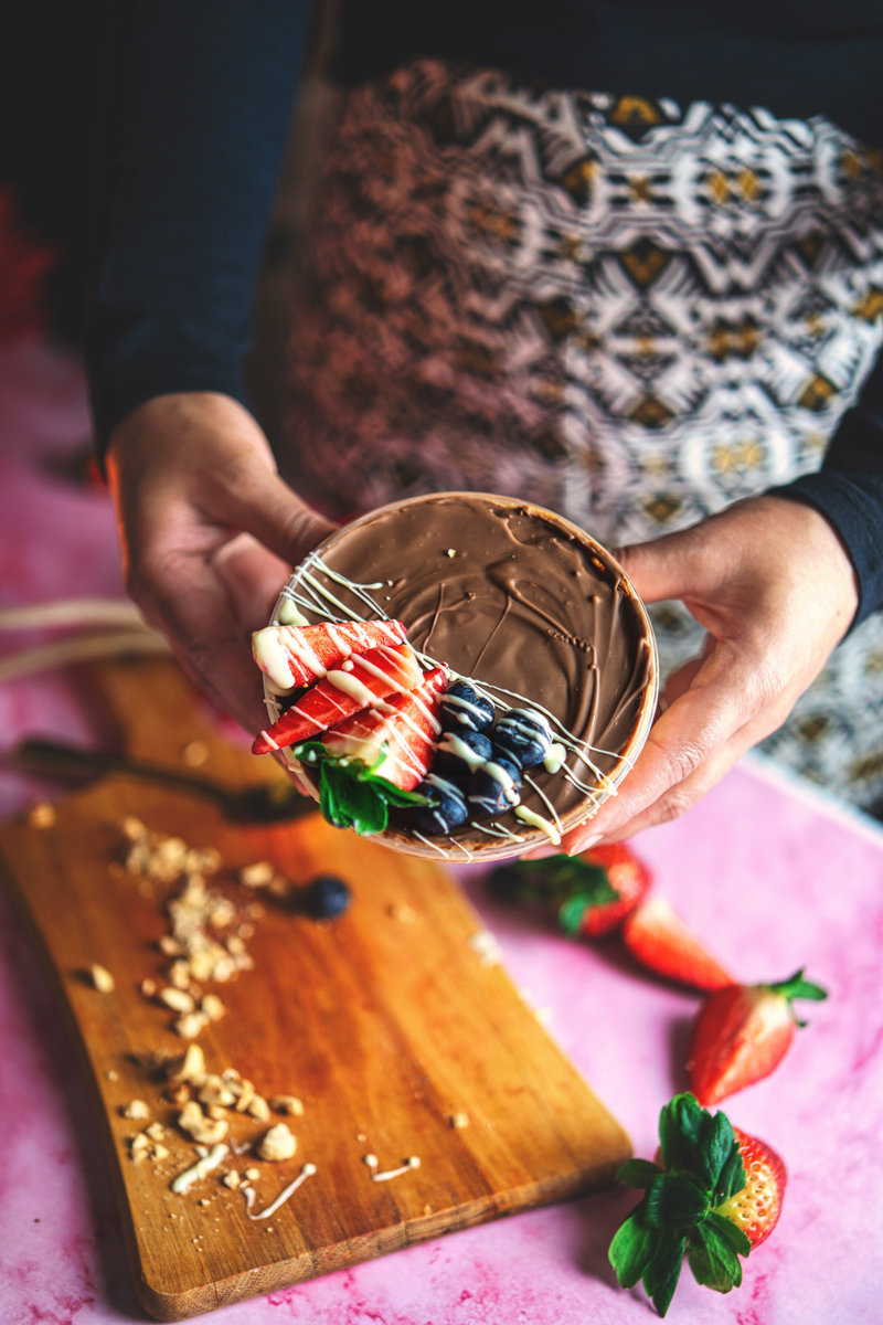 Hands presenting a peanut butter yogurt bowl garnished with sliced strawberries and blueberries drizzled with white and dark chocolate.