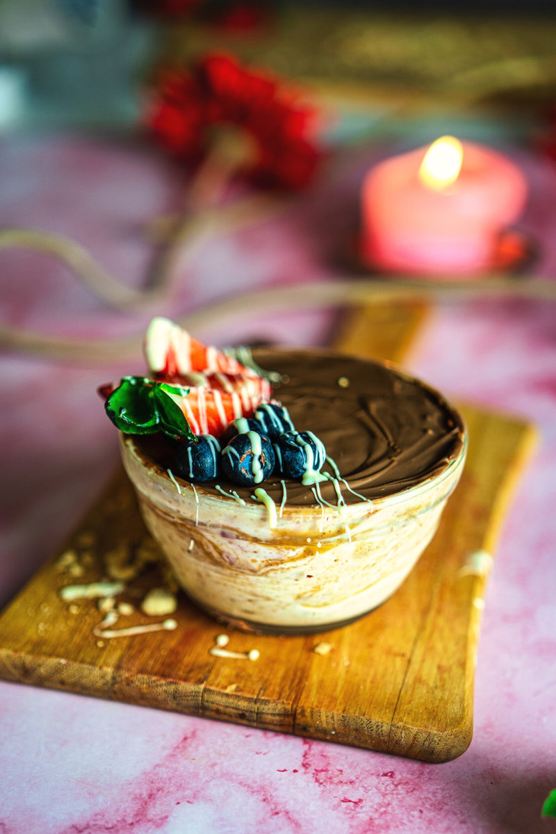 A peanut butter yogurt bowl with a smooth chocolate layer and a vibrant topping of strawberries and blueberries, on a pink surface next to a red flower and candlelight.