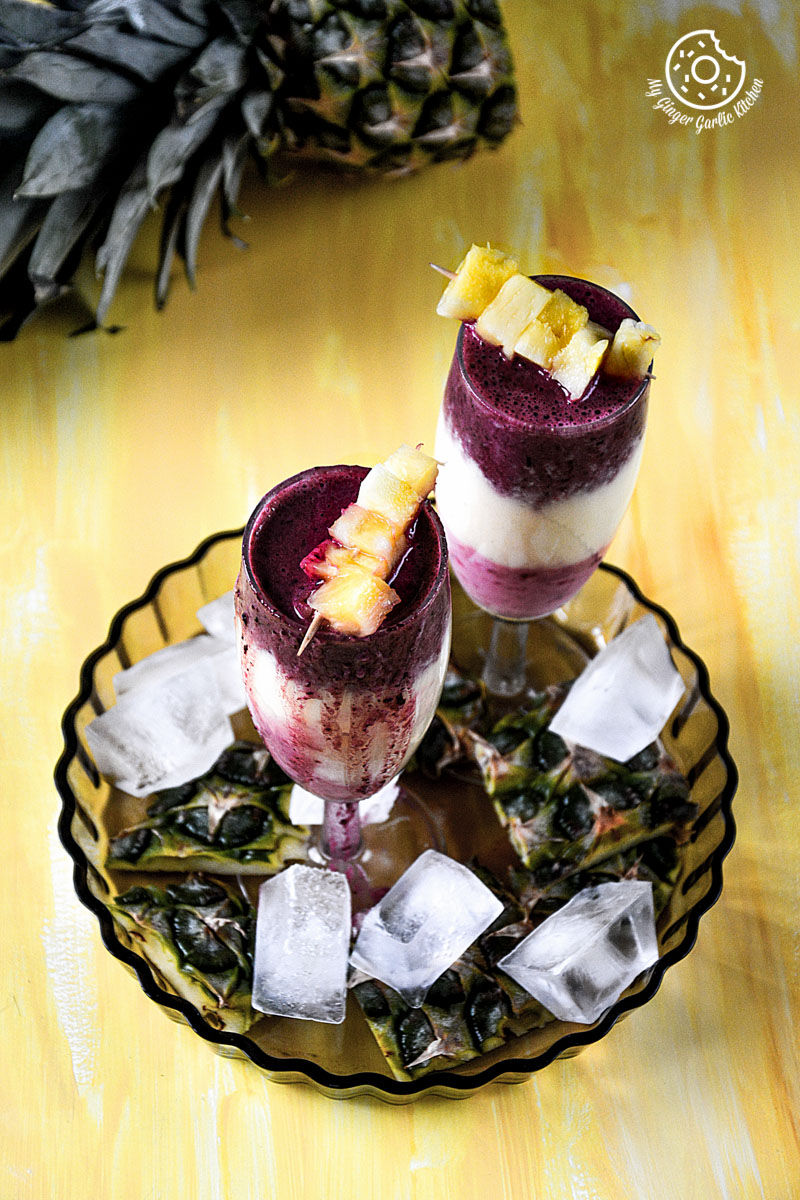 two glasses of pineapple blueberry raspberry breakfast layered smoothie and ice on a plate