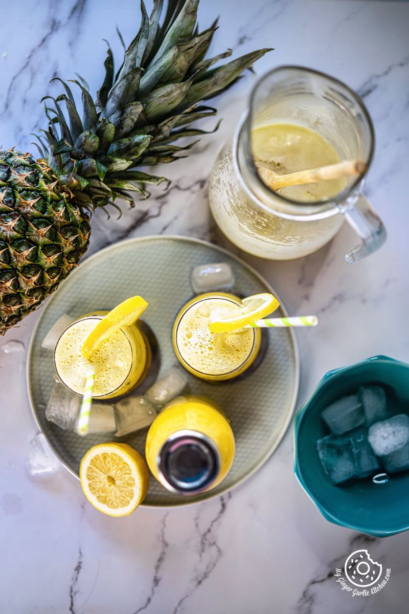 overhead shot of two pineapple juice glasses and a bottle in a plate and a pineapple and pitcher on the side